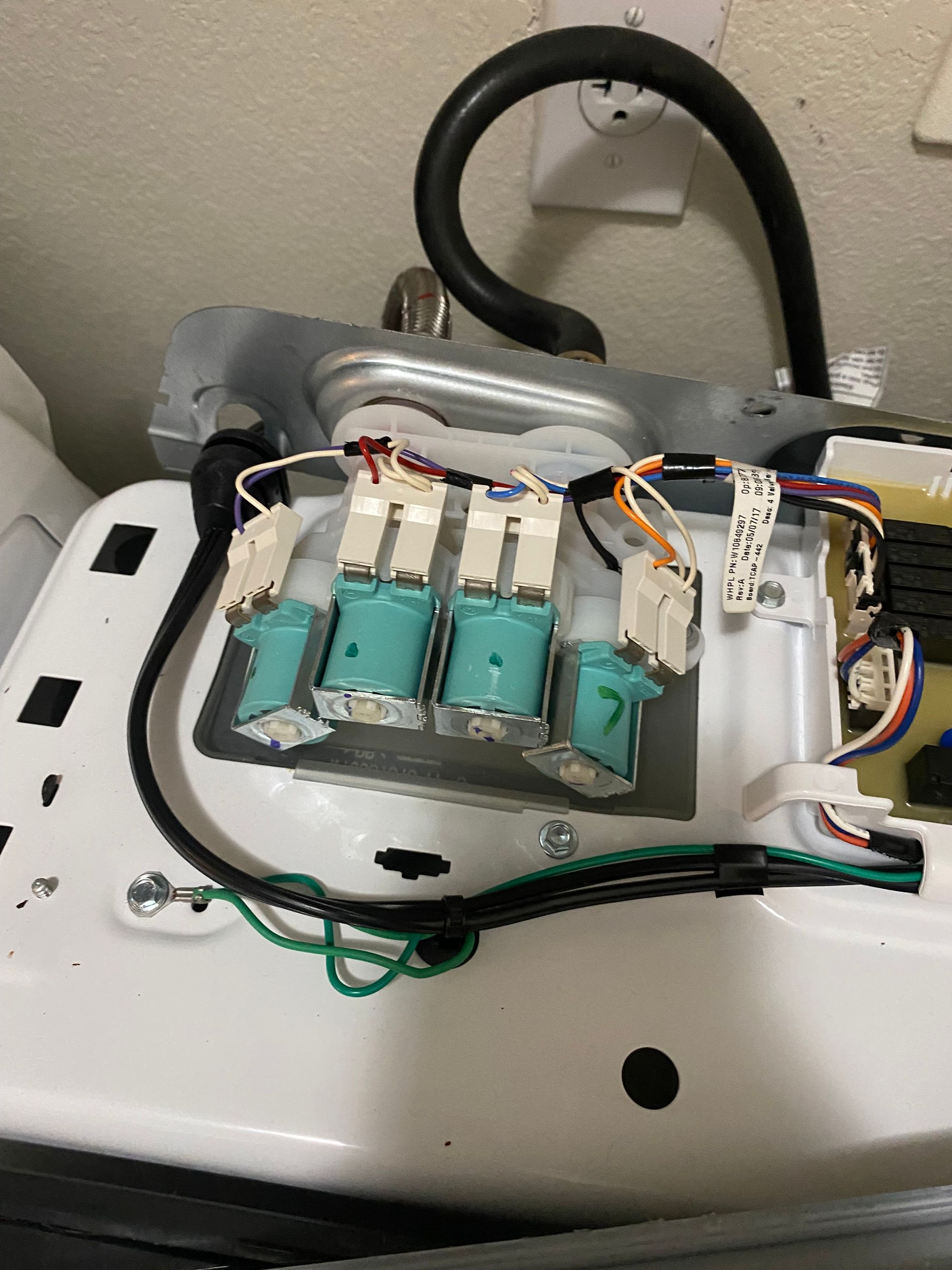 Top of a washing machine showing four blue water inlet valves, wiring, and a black hose.