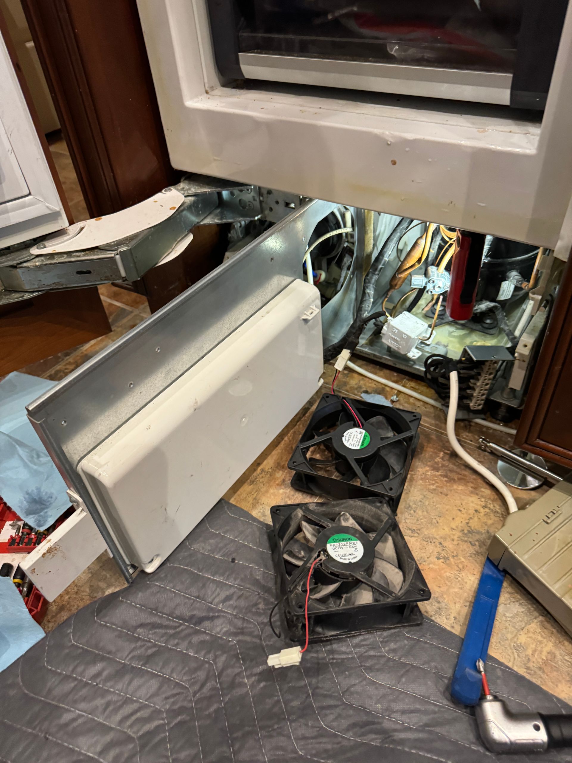Refrigerator repair: two fans, a metal cover, and exposed internal components under the appliance.