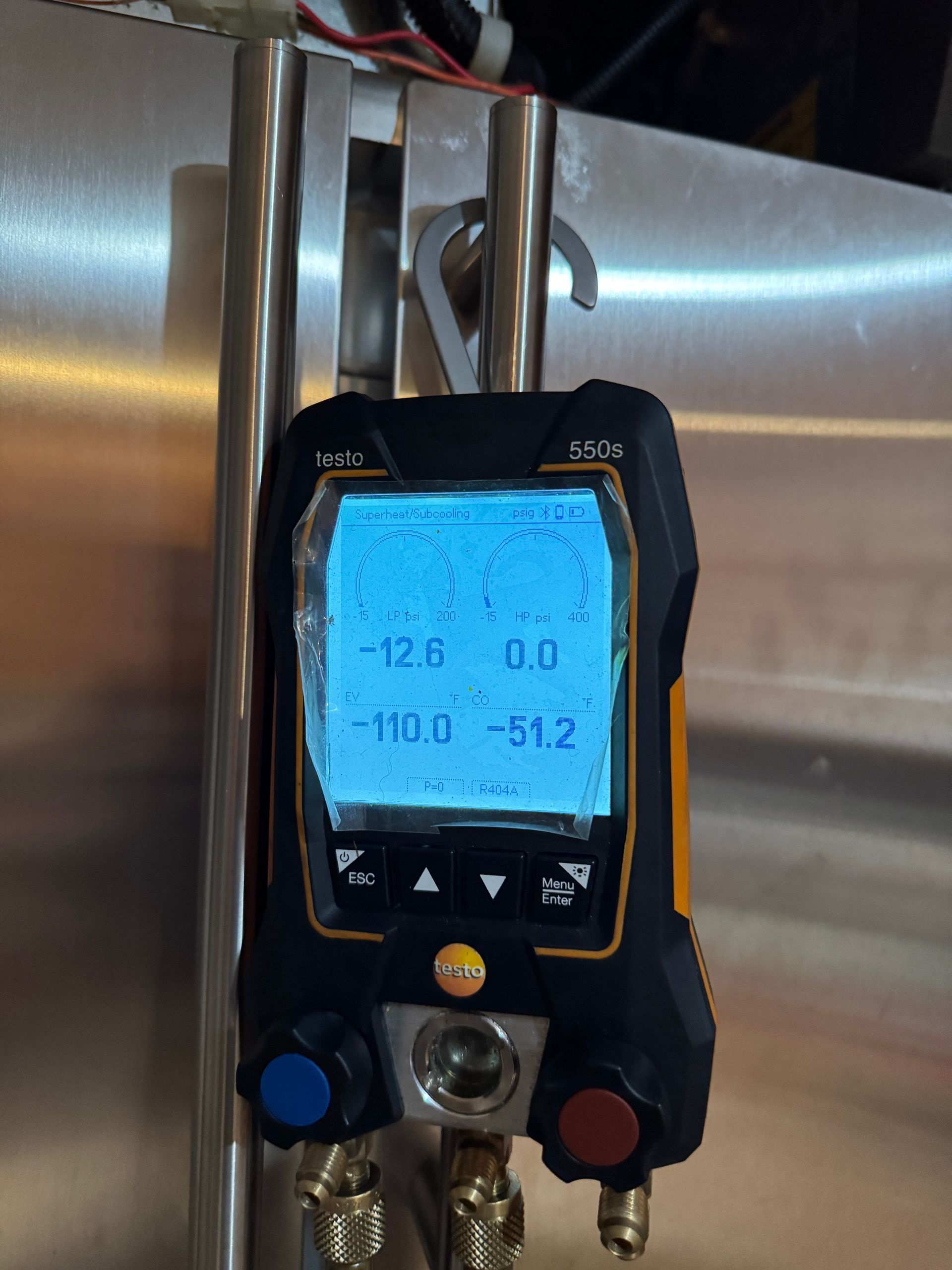 Digital refrigerant manifold gauge displaying pressure readings, hanging from a metal fixture.