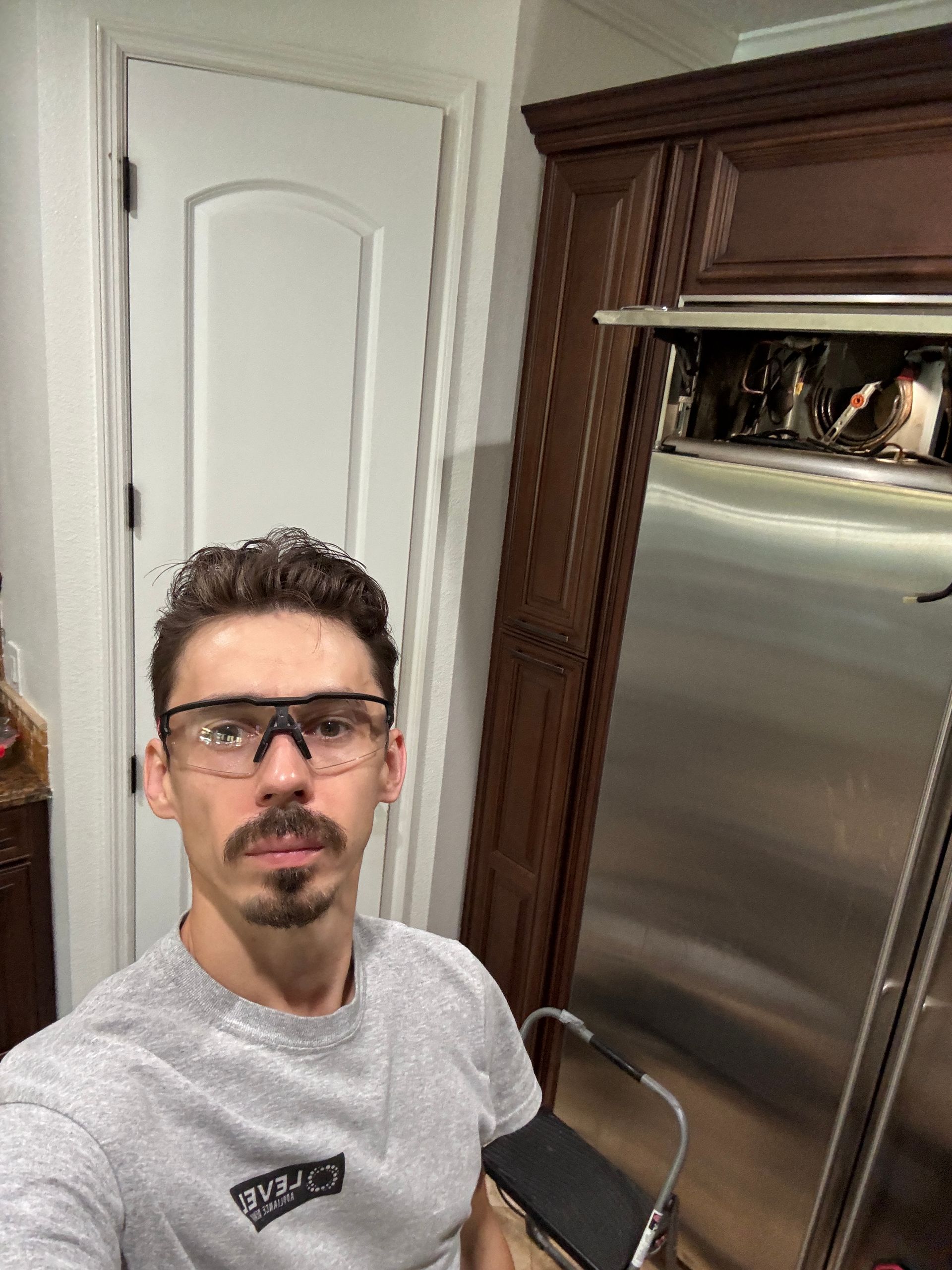Man wearing safety glasses stands near a stainless steel appliance, leaning on a small ladder in a kitchen.