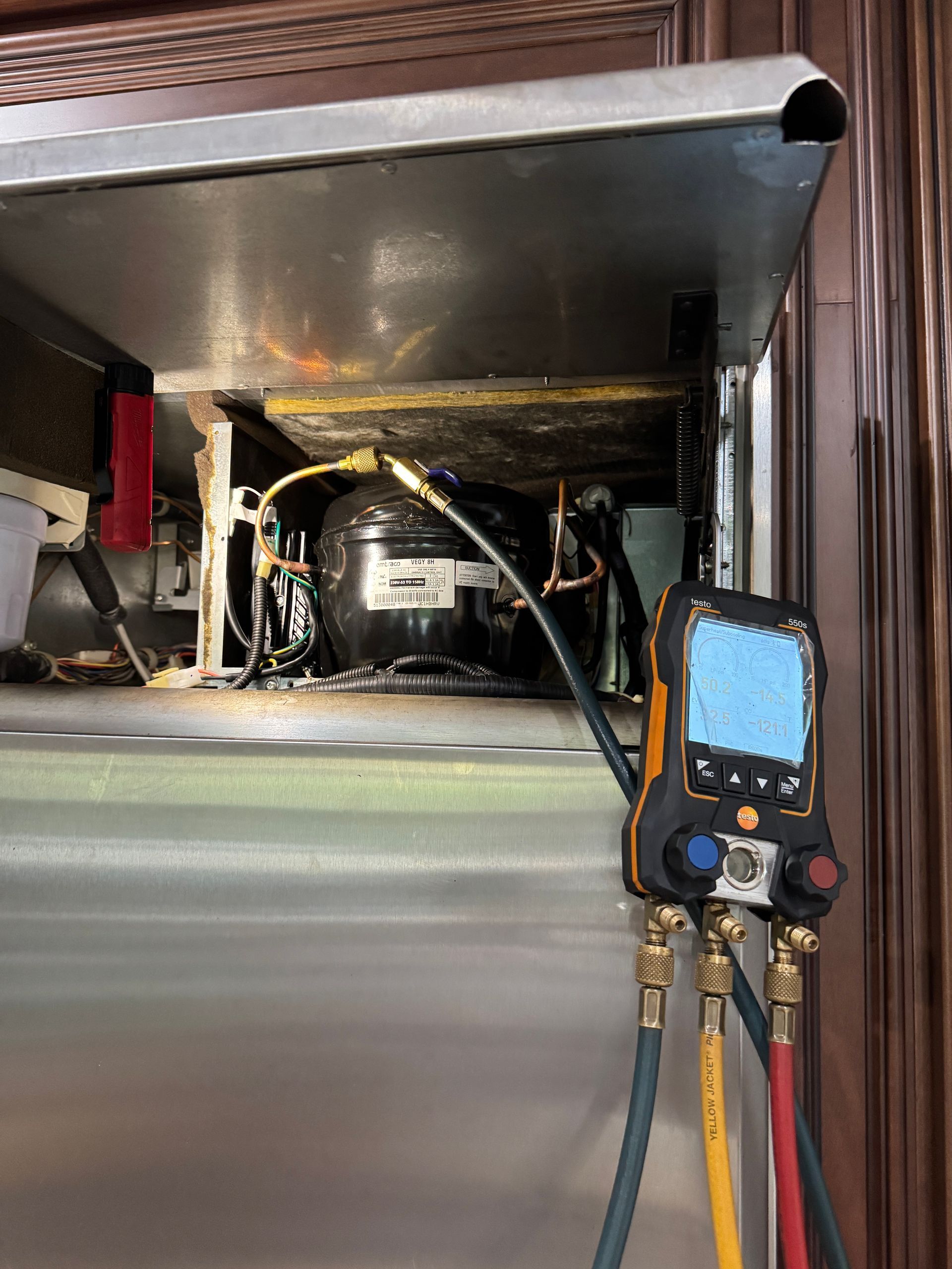Refrigeration unit being serviced. Digital gauge connected to hoses, assessing pressure. Stainless steel cabinet.