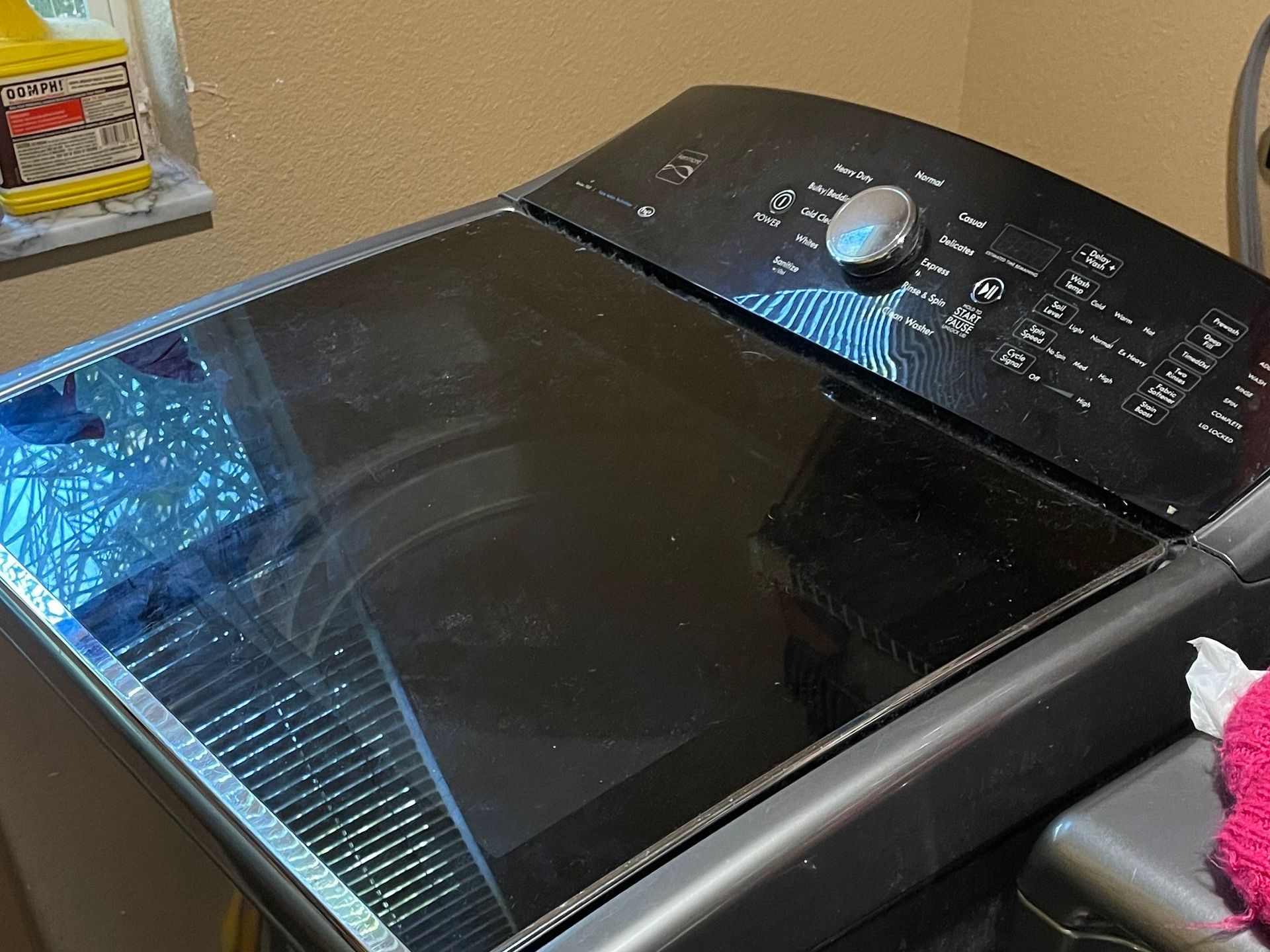 Black top-load washing machine with glass lid, control panel visible, in a laundry room.