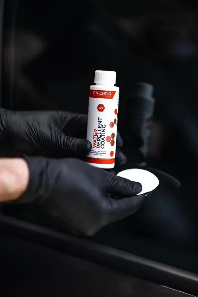 Gloved hands holding a small white-and-red bottle over a white pad in a dark car interior