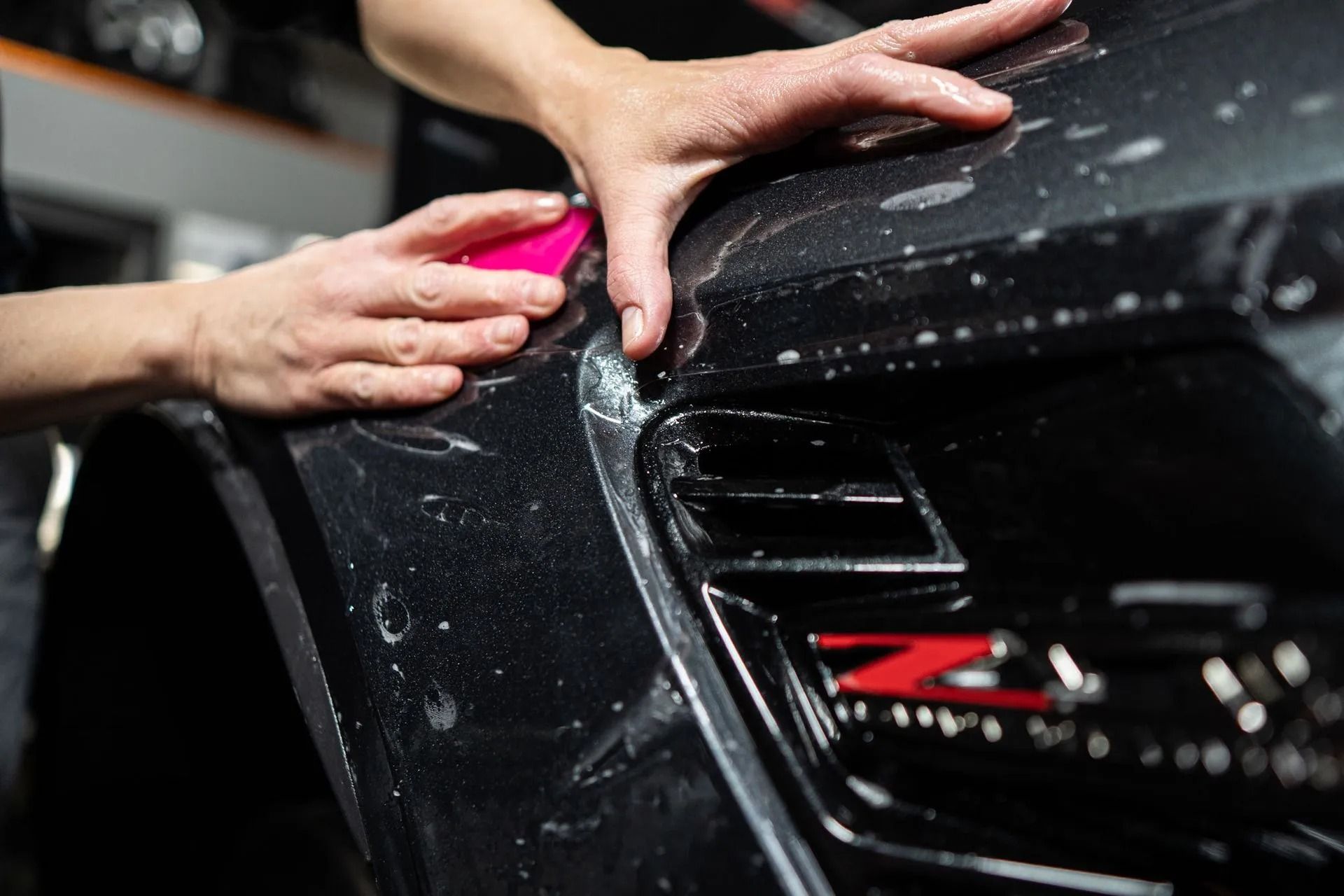 Hands cleaning a black car grille with a pink sponge, showing soap suds.