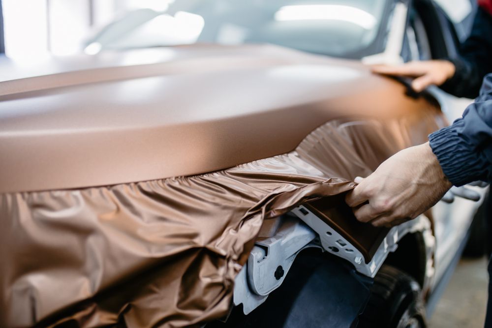 Car hood being wrapped with bronze vinyl by gloved hands in a workshop