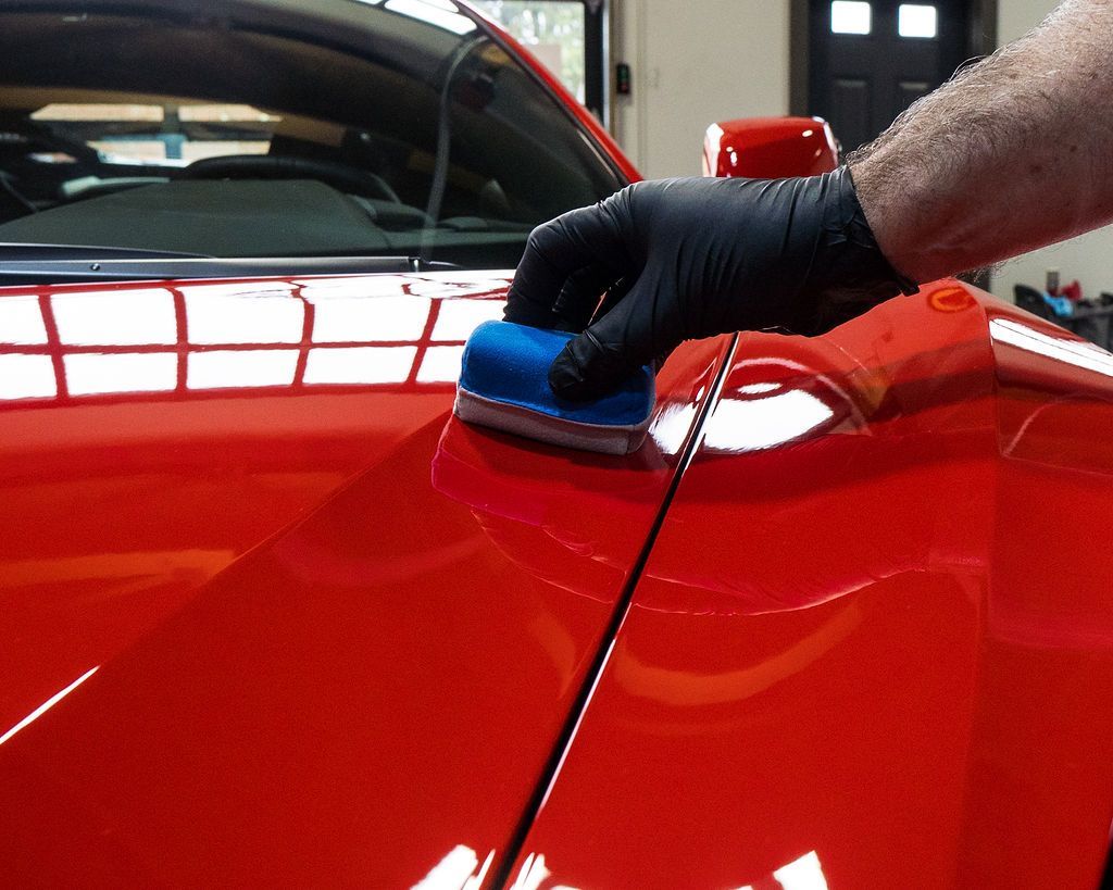 Gloved hand polishing a red car hood with a blue applicator in a garage