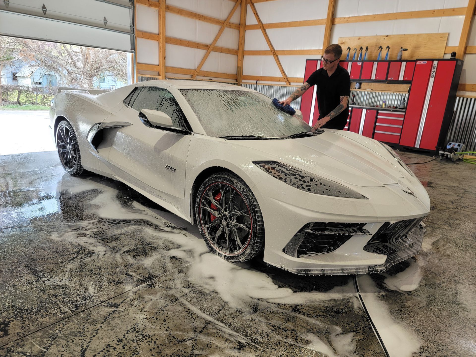 White sports car being washed in a garage, with foam on the floor and a person cleaning it.
