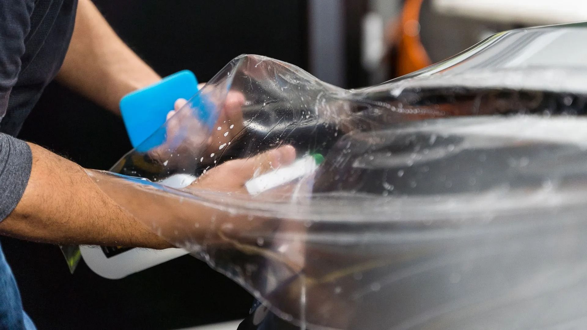 Hands smoothing clear plastic over a dark surface with a blue squeegee tool