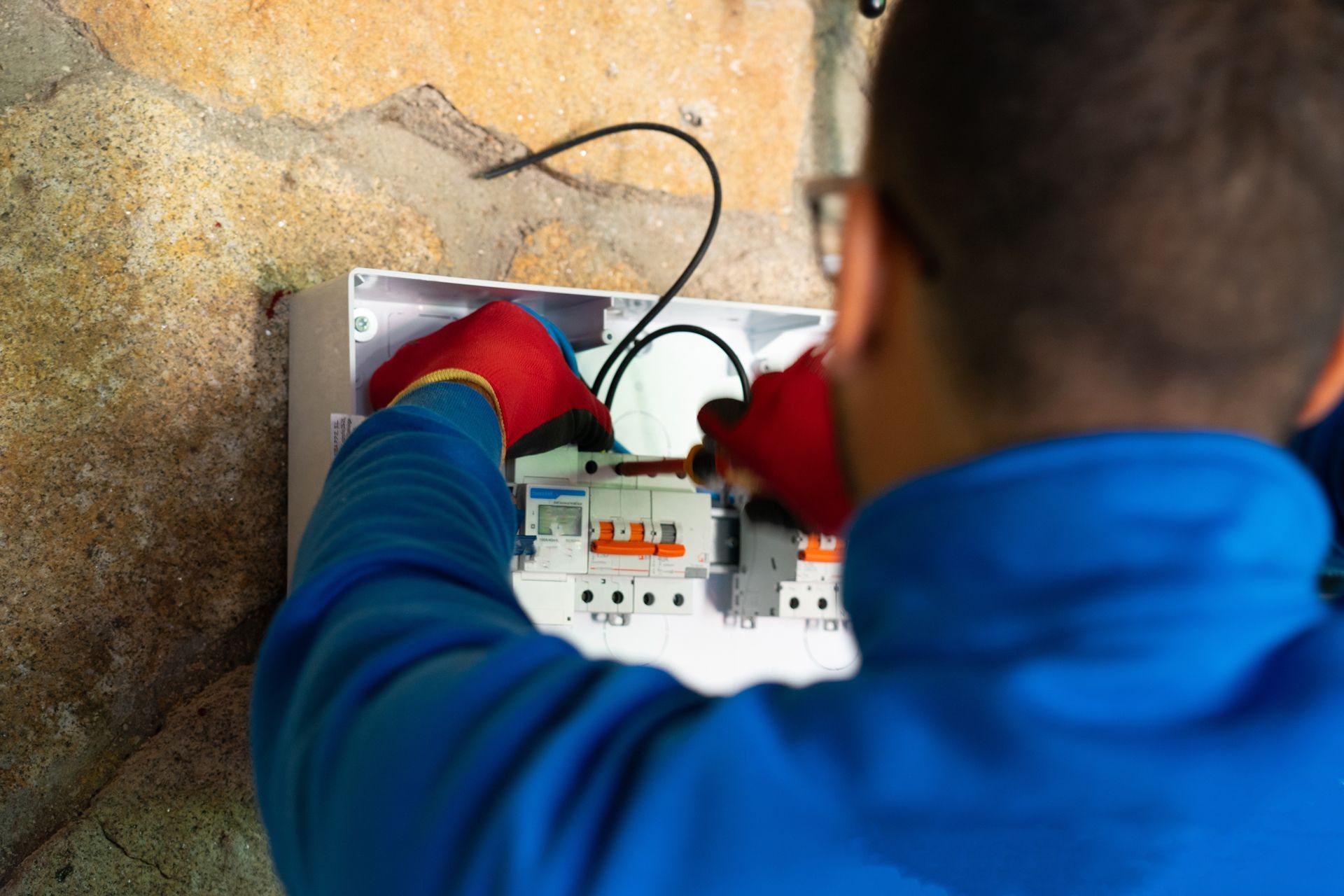 Electrician in blue jacket and red gloves wiring an open electrical panel on a wall