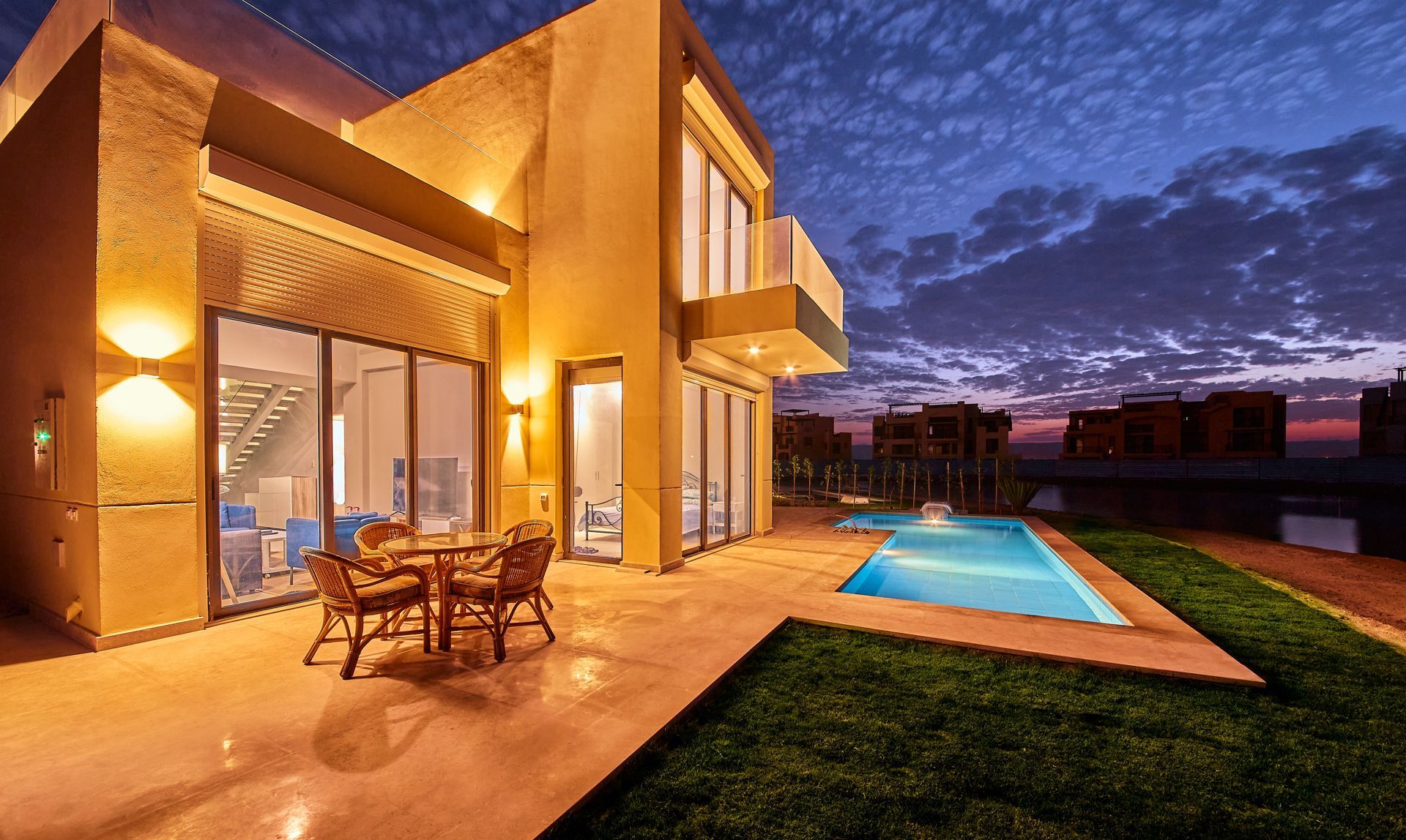 Modern house patio at dusk with lit interior, outdoor seating, and a narrow pool beside a grassy lawn.