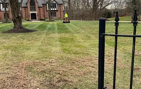 A grassy yard with a worker in a bright yellow vest near a brick building and black metal fence.