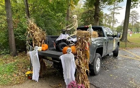 Decorated pickup truck with Halloween scarecrow and pumpkins parked outdoors by trees