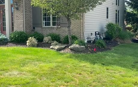 Corner of a brick-and-siding house with a green lawn, shrubs, rocks, and a small patio chair.