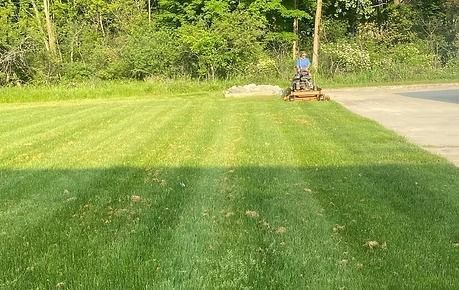 Lawn mower cutting grass near a driveway, with a green lawn and trees in the background