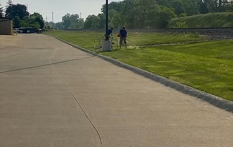 Wide concrete path beside a grassy field, with two people walking near a lamp post.