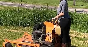 Person operating an orange mower in a grassy field, with a road and trees in the background