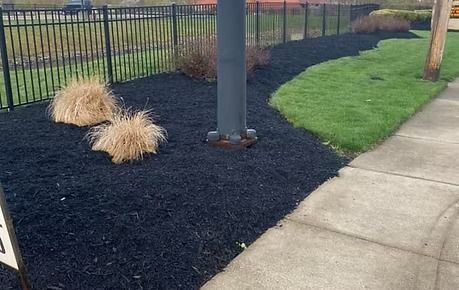 Black mulch around a playground post beside a sidewalk, with tan ornamental grasses and a metal fence.