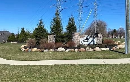 Landscaped entrance sign with rocks and shrubs beside a sidewalk on a sunny day