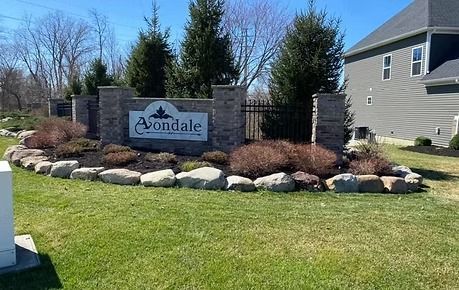 Stone community entrance sign reading “Avondale” with landscaping, grass, trees, and a house nearby