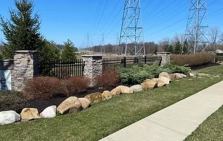 Landscaped roadside with boulders, shrubs, stone pillars, and a sidewalk beside power lines