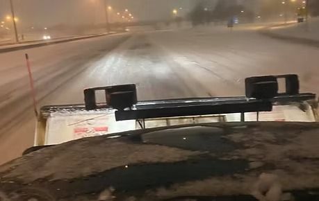 Snowy road seen from inside a vehicle at night, with plows and streetlights in the distance