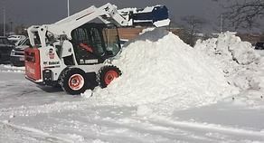 Small skid-steer loader pushing a large pile of snow across a snowy road.