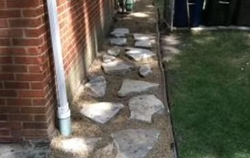 Narrow stone path beside a brick house, with stepping stones along the wall and grass on the right