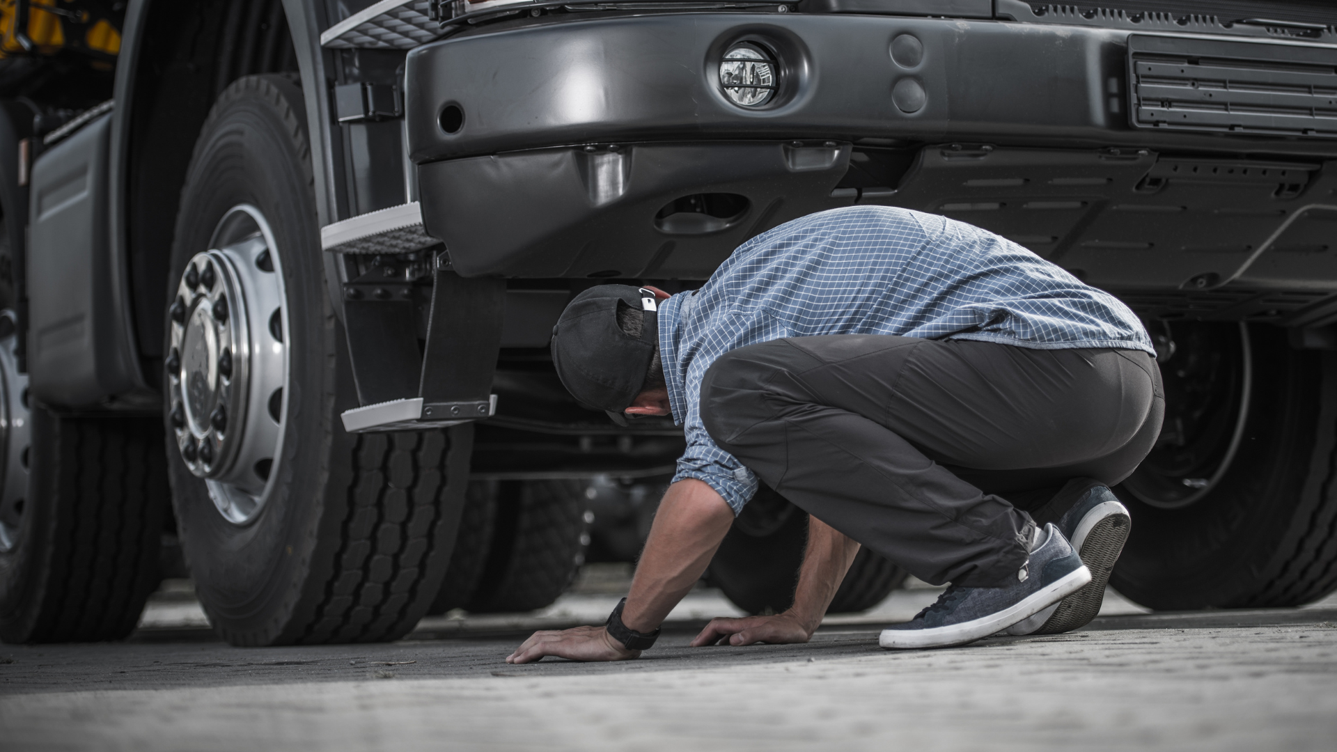 A person in a patterned shirt and dark pants crouches on the ground, inspecting the underside of a large black truck.