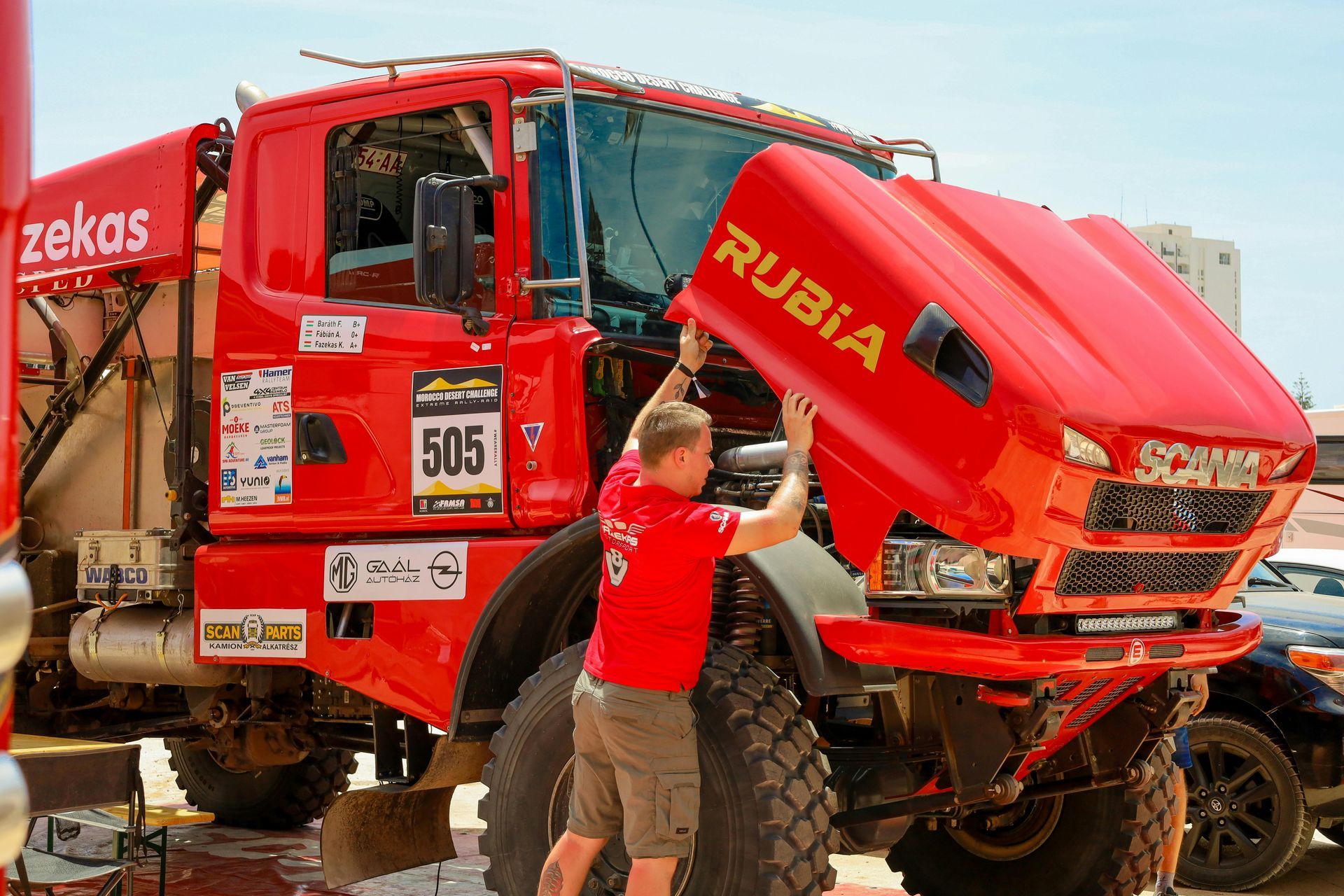 Red off-road race truck being serviced by a mechanic in a dusty outdoor pit area
