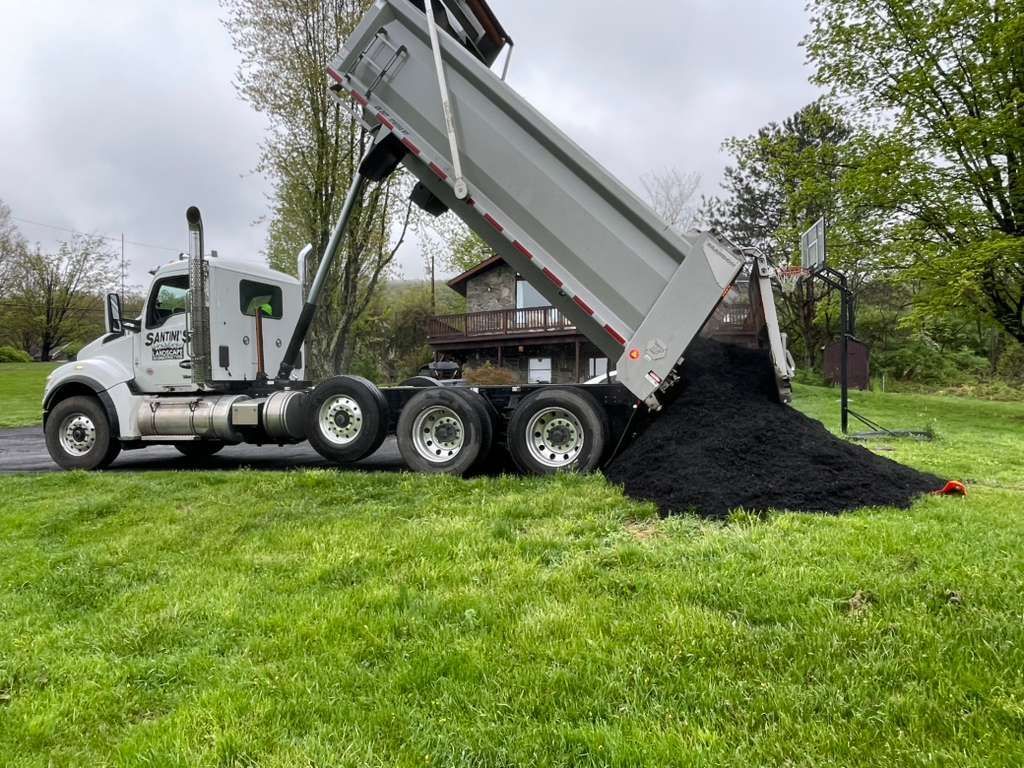 White dump truck unloading black gravel onto a grassy yard near a house.