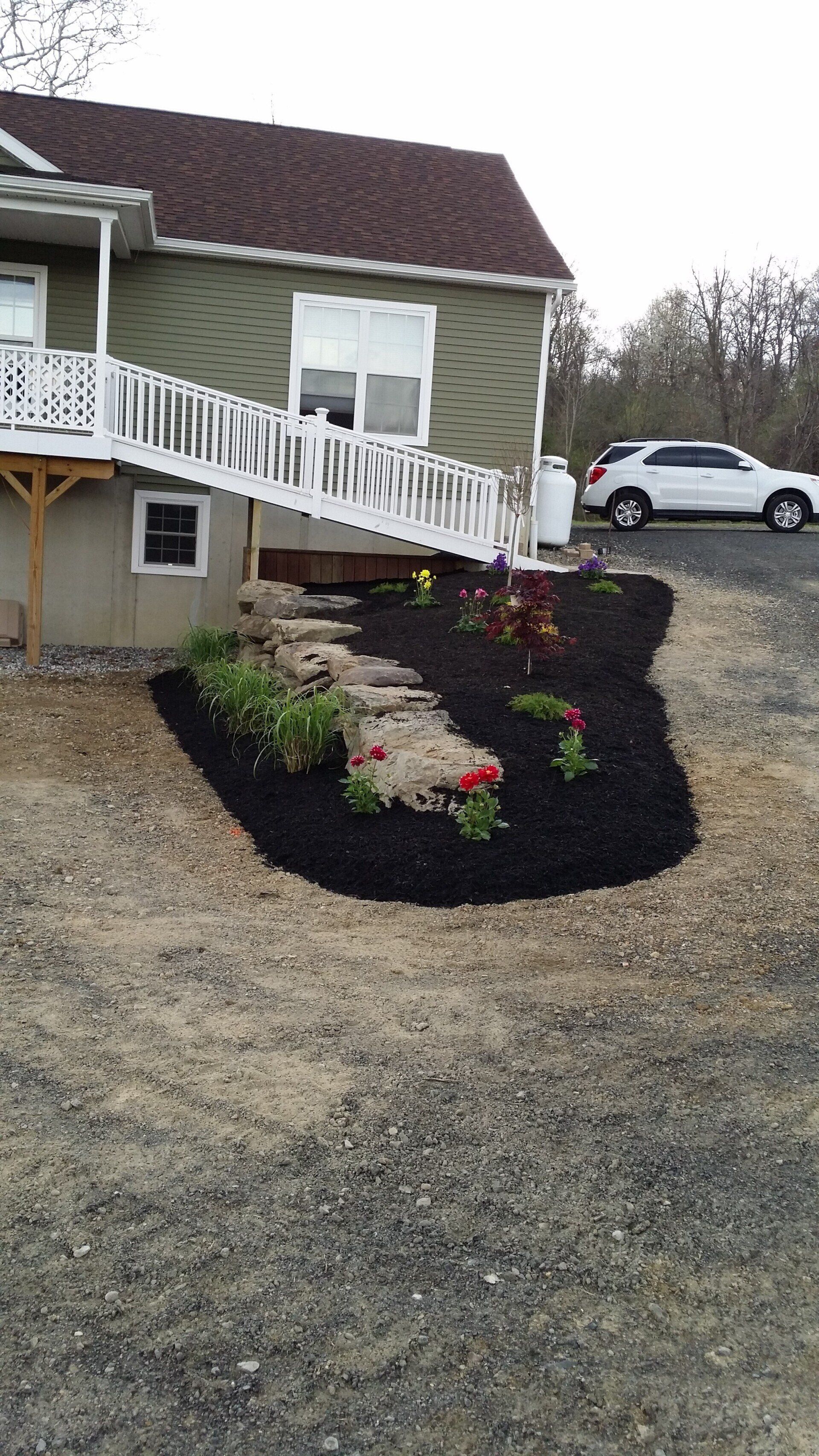 House with white staircase, gravel driveway, flower bed, and parked white car on a cloudy day