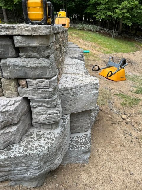 Stone retaining wall under construction with yellow tools on a dirt yard