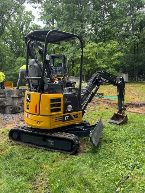 Yellow mini excavator on tracks digging on a grassy site with trees in the background.