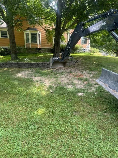 Excavator arm digging a hole in a grassy yard beside a house and trees