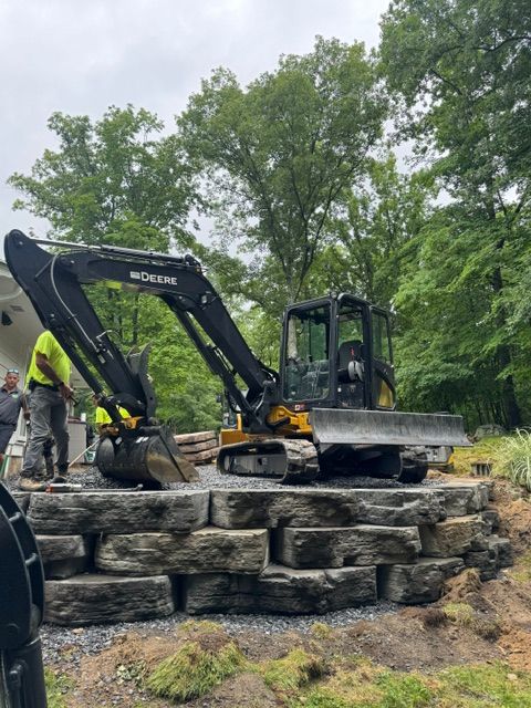 Excavator working on a stacked stone retaining wall outdoors with workers nearby and trees in the background