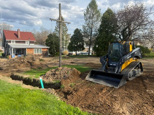 Backhoe excavator digging dirt beside a grassy yard with a house and utility pole nearby