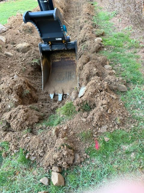 Excavator bucket digging a trench through grass and soil, with dirt piled along both sides.
