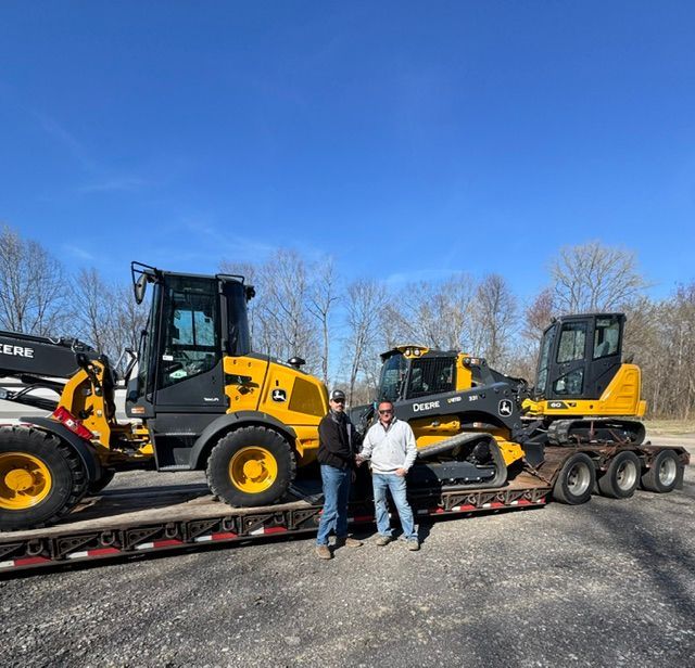 Two men standing in front of two yellow construction loaders on trailers  Steve Santini and John Deere salesman