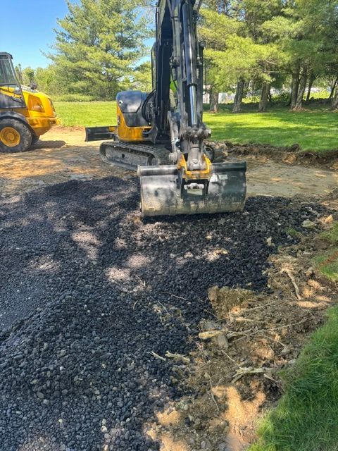 Excavator tamping dark gravel on a driveway beside a grassy yard and trees.
