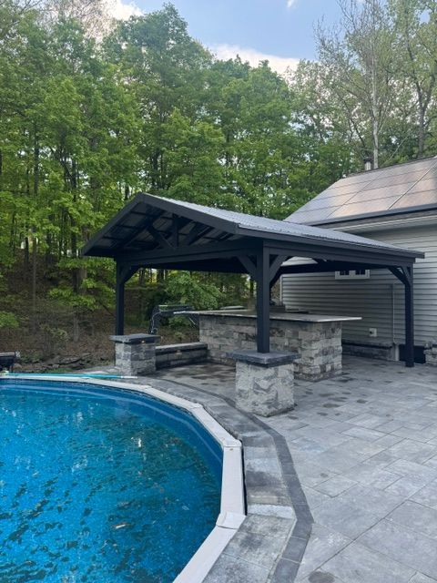 Backyard pool with stone bar and dark wood pavilion beside a house, surrounded by trees