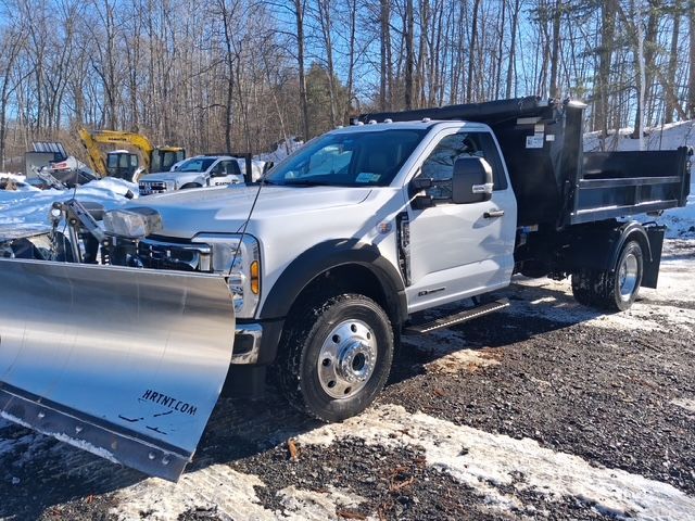 Santini's White dump truck with a front snow plow parked on a snowy lot with other vehicles and trees behind it