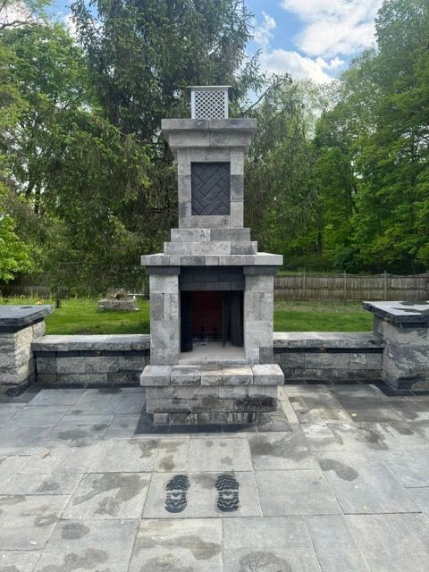 Camebridge Stone outdoor fireplace in a park, centered on a paved patio with trees in the background.