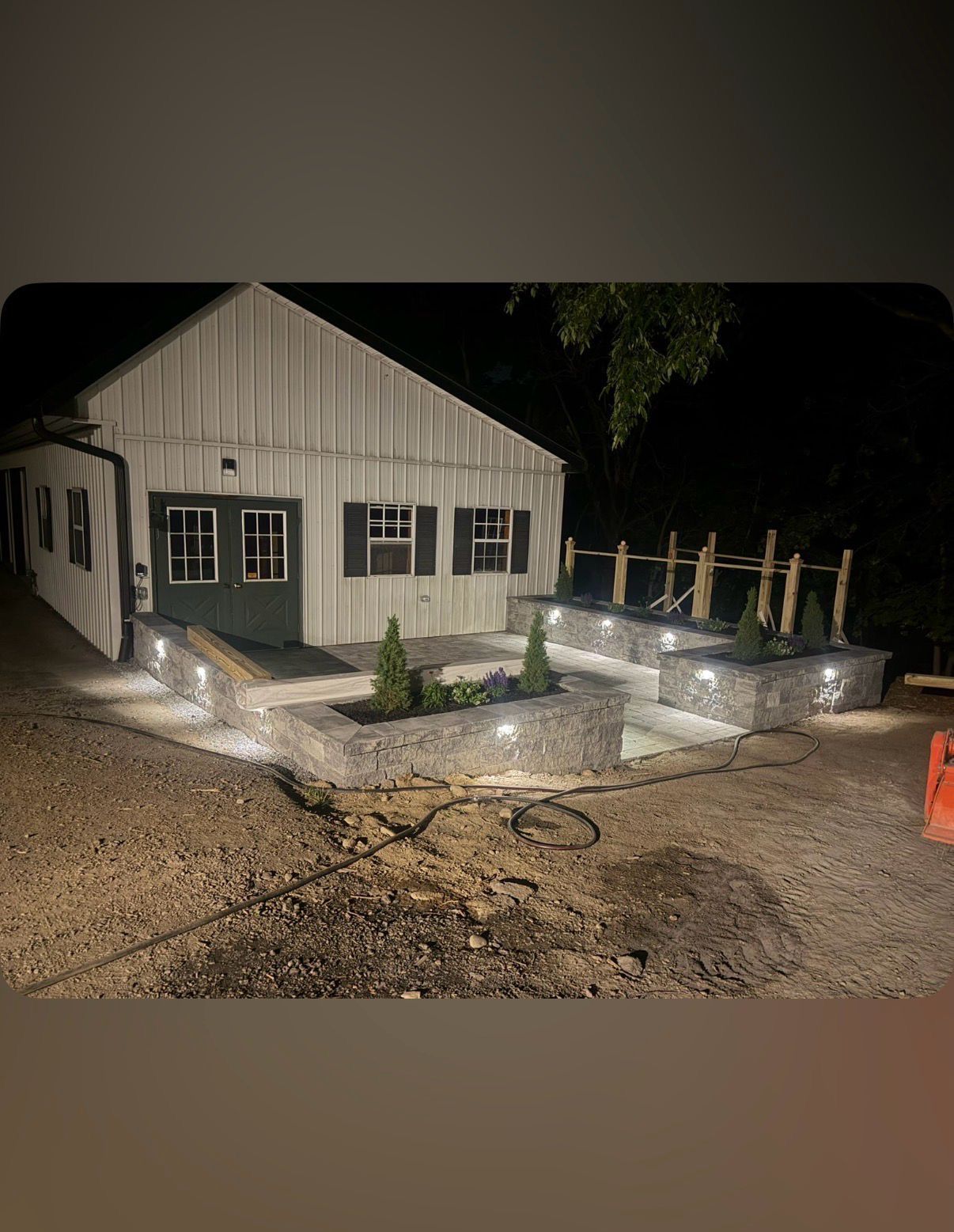 Small gray house at night with lit steps, gravel yard, and a wooden deck railing under construction