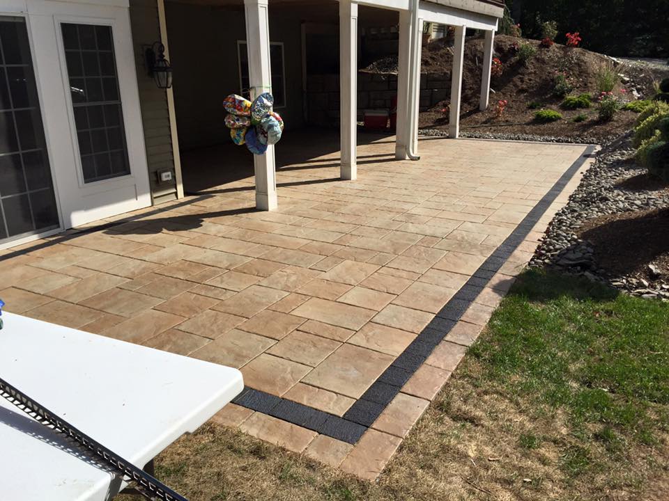 Back patio with tan pavers, dark border, and a grassy edge beside the house.