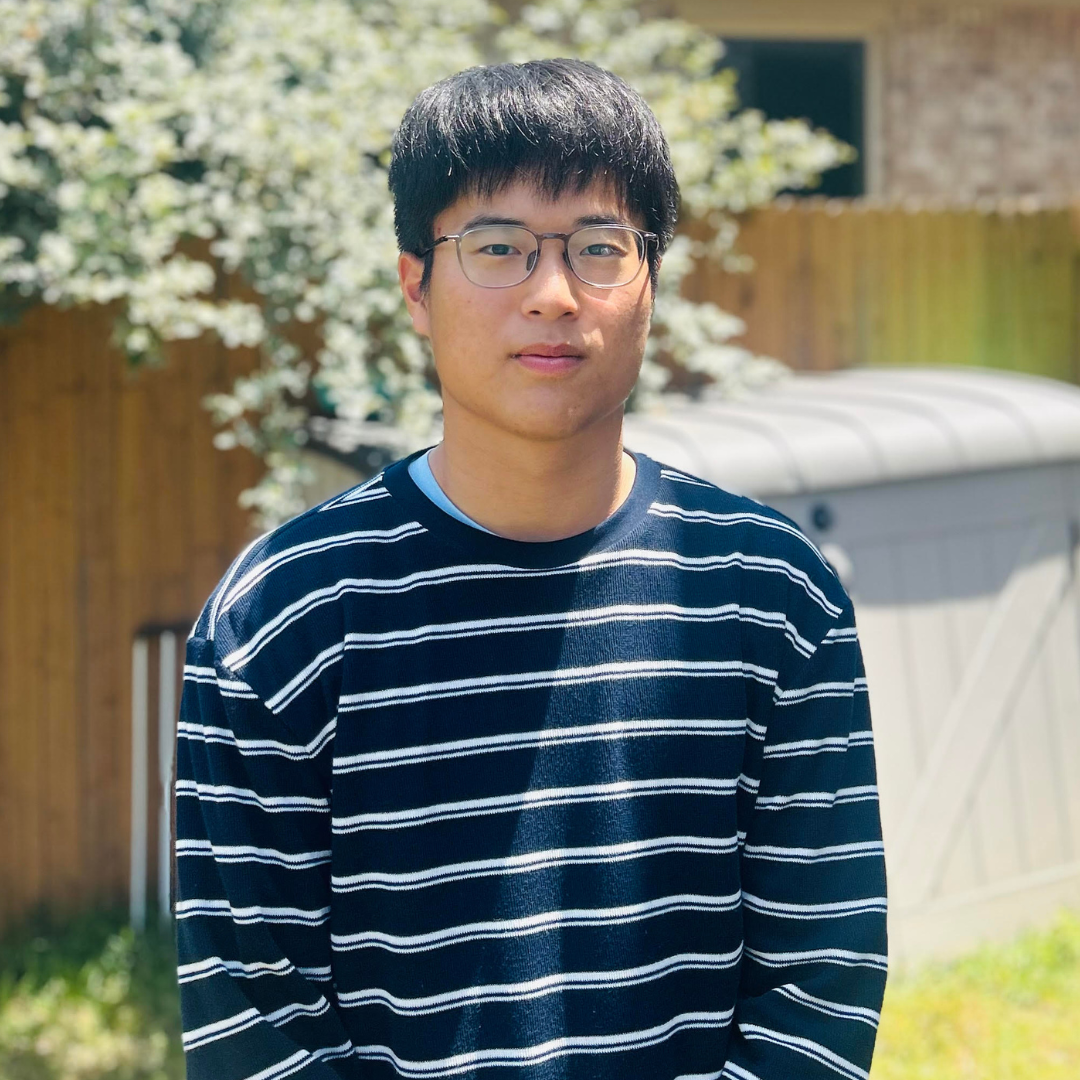 Person wearing glasses and a navy striped sweater stands outdoors in front of a wooden fence and shed.