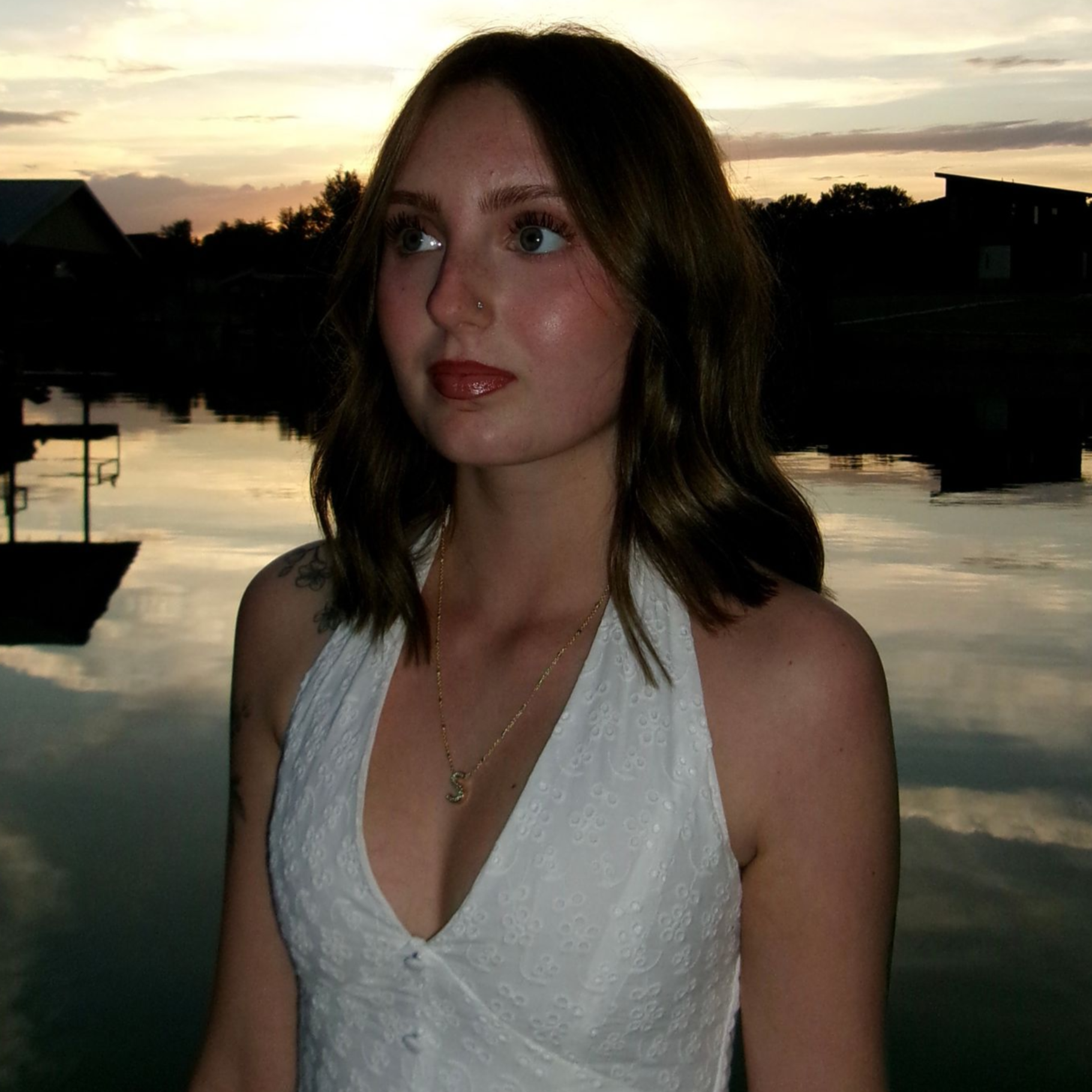 Woman in a white dress standing by water at sunset, with clouds and dark buildings in the background