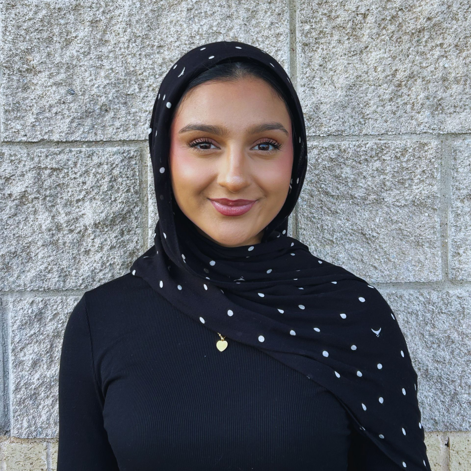 Person in a black polka-dot headscarf and top, smiling in front of a stone wall.
