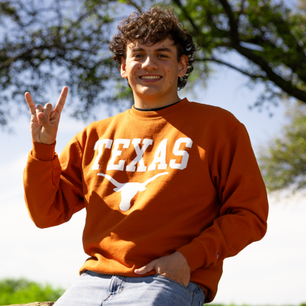 Person in an orange TEXAS sweatshirt making a hand sign outdoors on a sunny day