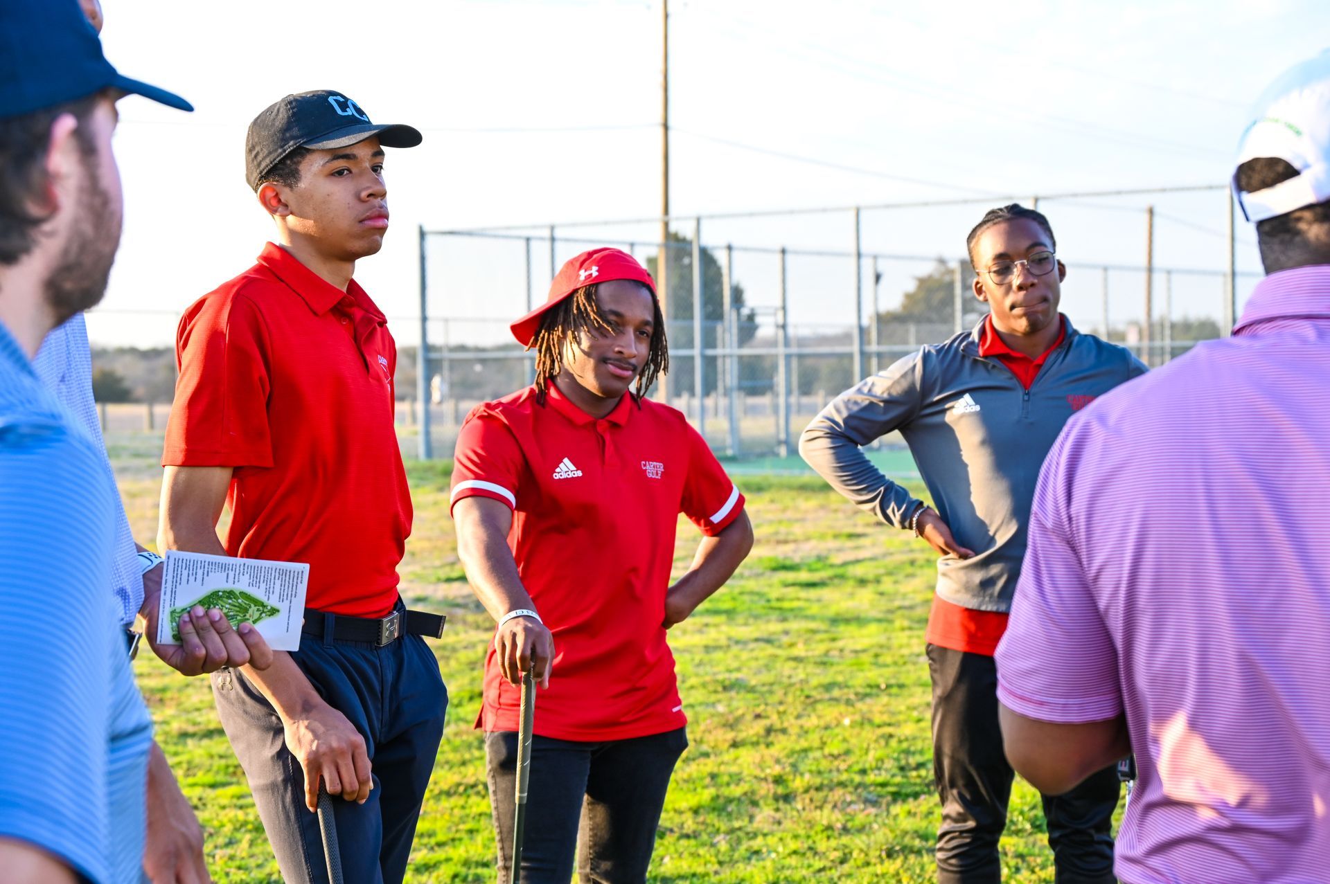 People in red shirts and a purple-striped shirt stand talking on a grassy sports field.