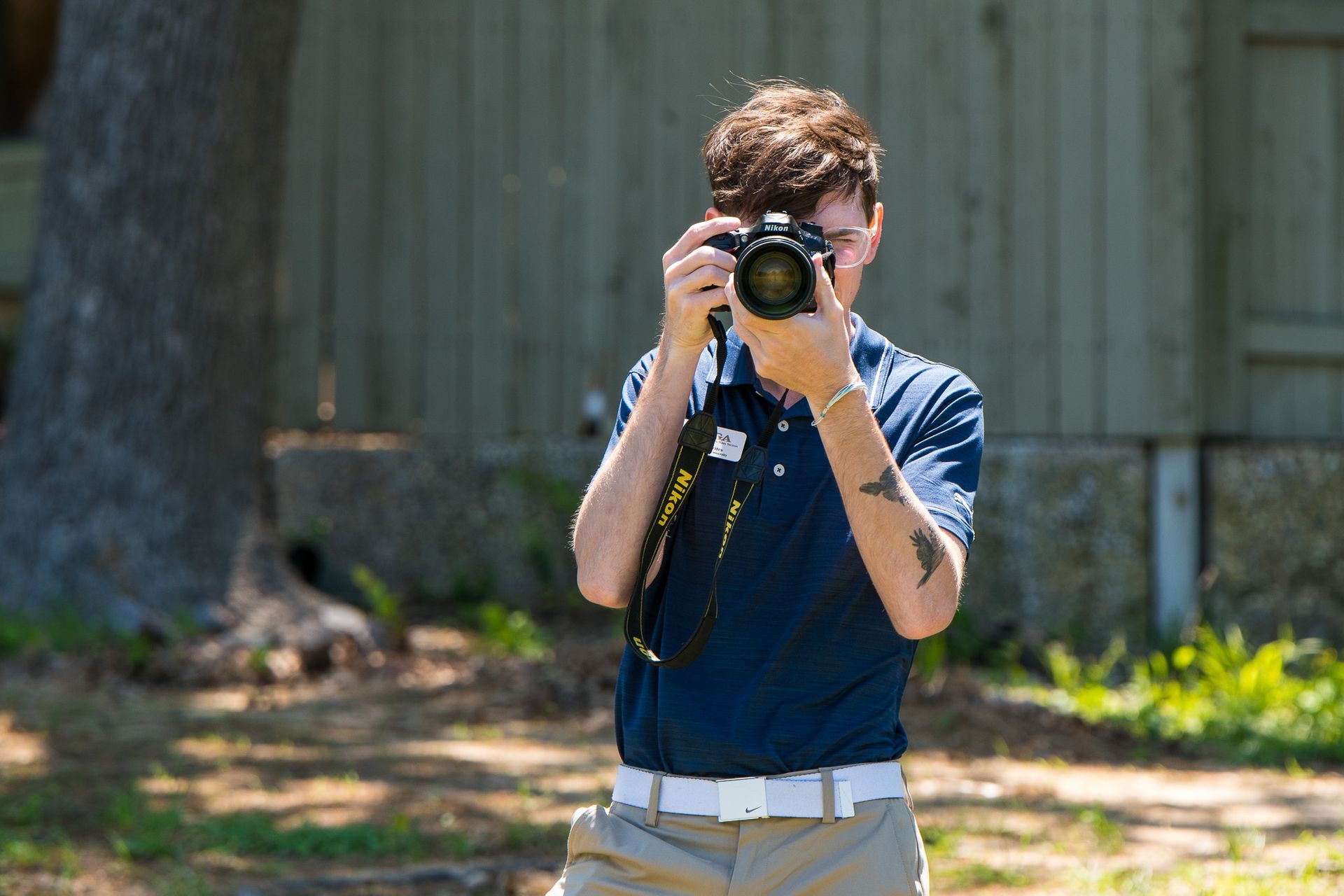 Person holding a camera outdoors in front of a wooden building and trees